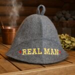 Pelēka filca pirts cepure ar izšūtu uzrakstu “REAL MAN” un sarkaniem zvaigžņu akcentiem, novietota uz koka lāvas pirts interjerā