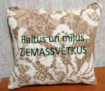 Aromātiskais spilventiņš ar uzrakstu "Baltus un mīļus ZIEMASSVĒTKUS" novietots uz koka virsmas. Attēlā redzams dekoratīvs auduma raksts un svētku vēlējums zaļā izšuvumā.