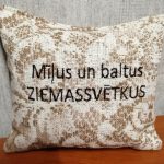 Aromātiskais spilventiņš ar uzrakstu "Mīļus un baltus ZIEMASSVĒTKUS" novietots uz koka virsmas. Attēlā redzams dekoratīvs auduma raksts un svētku vēlējums uz spilventiņa.