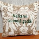 Aromātiskais spilventiņš ar uzrakstu "Veiksmi Jaunajā gadā!" novietots uz koka virsmas. Attēlā redzams dekoratīvs auduma raksts un izšūts svētku vēlējums.