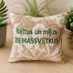 Aromātiskais auduma spilventiņš ar uzrakstu “Baltus un mīļus Ziemassvētkus”, novietots uz gaišas koka virsmas