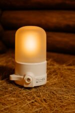Balta pirts lampa ar matētu kupolu, novietota uz salmiem koka pirts interjerā