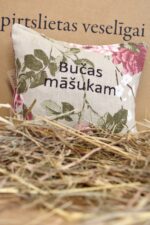 Aromātiskais spilventiņš ar uzrakstu "Bučas māšukam" uz salmu fona. Attēlā redzams dekoratīvs spilventiņš ar ziedu rakstu un sirsnīgu uzrakstu.