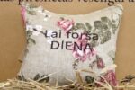Aromātiskais spilventiņš ar uzrakstu "Lai forša diena" tuvplānā uz salmu fona. Attēlā izcelta lina auduma tekstūra, ziedu raksts un lielais novēlējuma uzraksts.