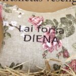 Aromātiskais spilventiņš ar uzrakstu "Lai forša diena" tuvplānā uz salmu fona. Attēlā izcelta lina auduma tekstūra, ziedu raksts un lielais novēlējuma uzraksts.