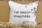 Aromātiskais spilventiņš ar uzrakstu "Lai sapņi piepildās" tuvplānā uz salmu fona. Attēlā izcelts spilventiņa auduma pinums, dekoratīvie ziedu elementi un labi salasāms novēlējuma uzraksts.
