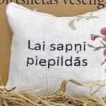 Aromātiskais spilventiņš ar uzrakstu "Lai sapņi piepildās" tuvplānā uz salmu fona. Attēlā izcelts spilventiņa auduma pinums, dekoratīvie ziedu elementi un labi salasāms novēlējuma uzraksts.