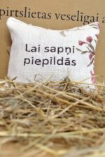 Aromātiskais spilventiņš ar uzrakstu "Lai sapņi piepildās" uz salmu fona. Attēlā redzams dekoratīvs spilventiņš ar ziedu motīviem un iedvesmojošu vēlējumu.