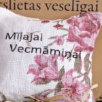 Aromātiskais spilventiņš ar uzrakstu "Mīļajai Vecmāmiņai" uz dekoratīva fona. Attēlā redzams spilventiņš ar rozā ziedu rakstu un sirsnīgu veltījumu vecmāmiņai.