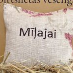 Aromātiskais spilventiņš ar uzrakstu "Mīļajai" tuvplānā uz salmu fona. Attēlā izcelta lina auduma tekstūra, dekoratīvie ziedu elementi un labi salasāms uzraksts.