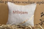 Aromātiskais spilventiņš ar uzrakstu "Mīļajam" tuvplānā uz dekoratīva fona. Attēlā izcelta lina auduma tekstūra, izšūtais uzraksts un spilventiņa forma.