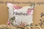 Aromātiskais spilventiņš ar uzrakstu "Paldies" tuvplānā uz dekoratīva fona. Attēlā izcelts spilventiņa auduma raksts, ziedu motīvi un izšūtais pateicības uzraksts.