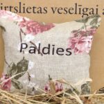 Aromātiskais spilventiņš ar uzrakstu "Paldies" tuvplānā uz dekoratīva fona. Attēlā izcelts spilventiņa auduma raksts, ziedu motīvi un izšūtais pateicības uzraksts.