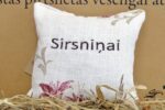 Aromātiskais spilventiņš ar uzrakstu "Sirsniņai" tuvplānā uz salmu fona. Attēlā izcelta spilventiņa faktūra, izšūtais uzraksts un dekoratīvie ziedu elementi stūros.