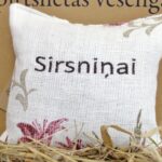 Aromātiskais spilventiņš ar uzrakstu "Sirsniņai" tuvplānā uz salmu fona. Attēlā izcelta spilventiņa faktūra, izšūtais uzraksts un dekoratīvie ziedu elementi stūros.