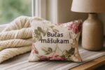 Aromātiskais auduma spilventiņš ar uzrakstu “Bučas māšukam”, novietots uz gaišas koka virsmas pie loga