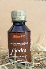 Gaisa aromatizētājs pirtīm un saunām "Ciedrs" uz salmu fona. Attēlā redzama tumša plastmasas pudele ar brūnu etiķeti un metālisku skrūvējamu vāciņu.