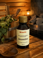 Gaisa aromatizētājs pirtīm ar jasmīna aromātu 120 ml tumšā stikla pudelē novietots uz koka galda pirts interjerā. Blakus redzams koka pirts spainis ar slotu, pirts krāsns akmeņi un aizdegta svece.