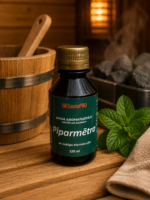 Gaisa aromatizētājs Piparmētra 120 ml tumšā pudelē novietots uz koka lāvas pirts interjerā. Blakus redzama svaiga piparmētra, dvielis un fonā – koka spainis un pirts krāsns.