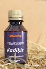 Gaisa aromatizētājs pirtīm un saunām "Kadiķis" uz salmu fona. Attēlā redzama tumša plastmasas pudele ar violetu etiķeti un metālisku skrūvējamu vāciņu.