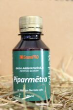 Gaisa aromatizētājs pirtīm un saunām "Piparmētra" uz salmu fona. Attēlā redzama tumša plastmasas pudele ar zaļu etiķeti un skrūvējamu vāciņu.
