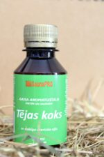Gaisa aromatizētājs pirtīm un saunām "Tējas koks" uz salmu fona. Attēlā redzama tumša plastmasas pudele ar zaļu etiķeti un metālisku skrūvējamu vāciņu.