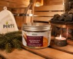 Burciņa ar pirts jūras sāli "Kleopatras receptes" novietota siltā pirts atmosfērā uz koka lāvas. Attēlā redzams produkts kopā ar egļu zariem, aizdegtu sveci, pirts cepuri, koka ķipīti un krāsns akmeņiem fonā.