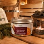 Burciņa ar pirts jūras sāli "Kleopatras receptes" novietota siltā pirts atmosfērā uz koka lāvas. Attēlā redzams produkts kopā ar egļu zariem, aizdegtu sveci, pirts cepuri, koka ķipīti un krāsns akmeņiem fonā.