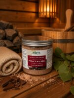Burciņa ar pirts sāli "Piena šokolāde" novietota siltā pirts atmosfērā uz koka lāvas. Attēlā redzams produkts kopā ar sarullētu dvieli, šokolādes gabaliņiem, jūras sāli un bērza lapām.
