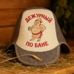 Divkrāsu filca pirts cepure ar krievu uzrakstu “ДЕЖУРНЫЙ ПО БАНЕ” un zīmējumu, novietota uz koka lāvas pirts interjerā
