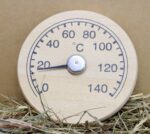 Apaļš koka pirts termometrs ar skalu līdz 140 °C, novietots uz salmiem, redzama termometra priekšpuse un temperatūras rādītājs