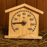 Koka pirts termometrs mājiņas formā ar temperatūras skalu līdz 140 °C, novietots uz salmu klājuma pirts vidē