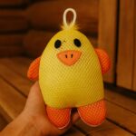 Bērnu pirts švamme pīlēna formā, turēta rokā pirts telpā uz koka sola