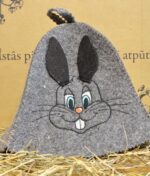 Pelēka pirts cepure ar lielu zaķa sejas izšuvumu priekšpusē, novietota uz salmiem, izcelts rotaļīgais dizains un filca faktūra