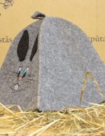 Pelēka pirts cepure ar izšūtu zaķa motīvu sānskatā, novietota uz salmiem, redzama cepures forma un izšuvuma detaļas
