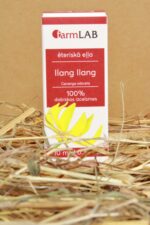 FarmLAB ilang ilang ēteriskā eļļa iepakojumā novietota uz salmiem, redzams produkta iepakojums ar ilang ilang ziedu attēlu