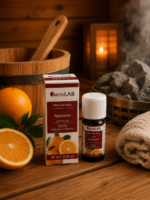 Ēteriskā eļļa Apelsīns 10 ml pudelītē ar iepakojumu novietota uz koka lāvas pirts interjerā. Blakus redzams svaigs apelsīns, fonā – koka spainis, dvielis un pirts krāsns.