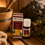 Ēteriskā eļļa Citrons 10 ml pudelītē ar iepakojumu novietota uz koka lāvas pirts interjerā. Fonā redzama pirts krāsns ar akmeņiem, koka spainis un pirtsslota.