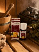 Ēteriskā eļļa Citrons 10 ml pudelītē ar iepakojumu novietota uz koka lāvas pirts interjerā. Fonā redzama pirts krāsns ar akmeņiem, koka spainis un pirtsslota.
