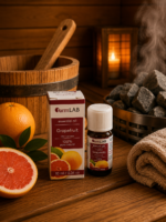 Ēteriskā eļļa Greipfrūts 10 ml pudelītē ar iepakojumu novietota uz koka lāvas pirts interjerā. Blakus redzams greipfrūts, fonā – koka spainis, dvielis un pirts krāsns.