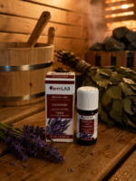 Ēteriskā eļļa Lavanda 10 ml pudelītē ar iepakojumu novietota uz koka lāvas pirts interjerā. Fonā redzama pirts slota, koka spainis un lavandas ziedi.