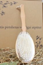 Ovāla koka masāžas birste ar gariem baltiem sariem un ērtu rokturi novietota uz salmu klājuma pie dekoratīva fona