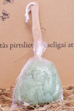 Gaiši zaļš pirts sūklis ar koka rokturi caurspīdīgā iepakojumā novietots uz salmu klājuma