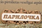 Koka dekoratīva pirts izkārtne ar uzrakstu “Парилочка”, novietota uz salmiem gaišā fonā
