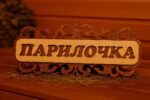 Koka dekoratīva pirts izkārtne ar uzrakstu “Парилочка”, novietota uz lāvas