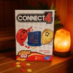 Galda spēles kaste “Connect 4 Grab & Go” uz koka lāvas pirts telpā, priekšplānā redzami spēles kauliņi, fonā sāls lampa