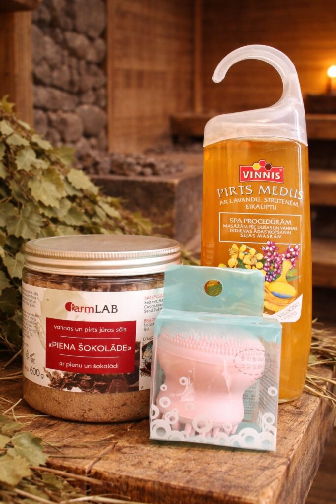 Pirts dāvanu komplekts ar pirts medu, vannas sāli un kopšanas līdzekli, novietots uz koka virsmas pirts interjerā