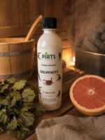 Pirts aromatizators ar greipfrūta aromātu 500 ml baltā pudelē novietots uz koka lāvas pirts interjerā. Priekšplānā redzama pārgriezta greipfrūta puse un pirts slota, fonā pirts krāsns ar akmeņiem.