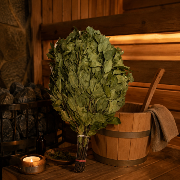 Ievas pirts slota novietota uz koka lāvas tradicionālā pirts interjerā. Blakus redzams koka ķipis, pirts akmeņi, degoša svece un trauks ar kaltētām Ievas lapām.