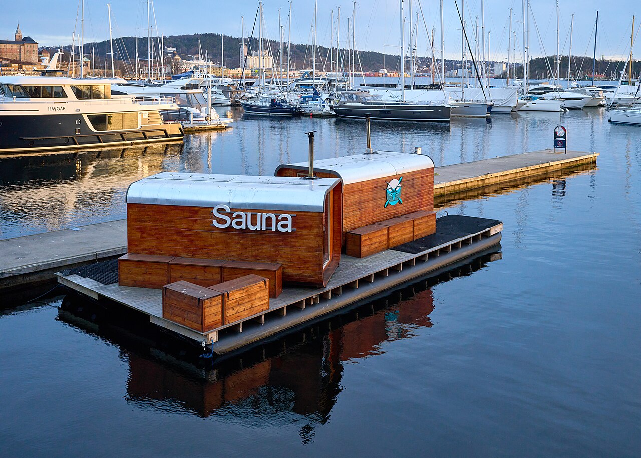 Peldoša koka sauna Oslo ostā ar jahtām un buru laivām fonā.